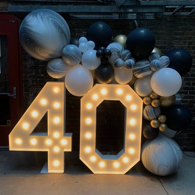 in a dimly lit room are large light up marquee numbers with a balloon garland of black silver white and marble balloons draped over