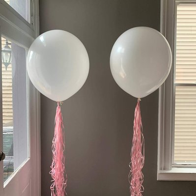 In front of a grey wall between windows are two white helium balloons with pink ribbons