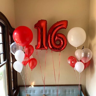 in front of a beige wall on a blue couch are two helium balloon bouquets of red clear and white balloons with two red mylar number balloons in between
