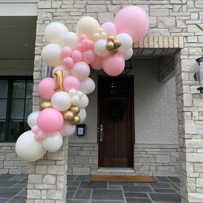 Over a stone entranceway to a home is a pink and white balloon garland with small gold accents and a gold mylar balloon number 1