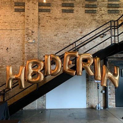 over a staircase in front of a brick wall are large gold mylar balloon letters for a birthday
