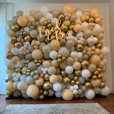 in front of a white wall is a large balloon wall of gold tan and white balloons for a bridal shower