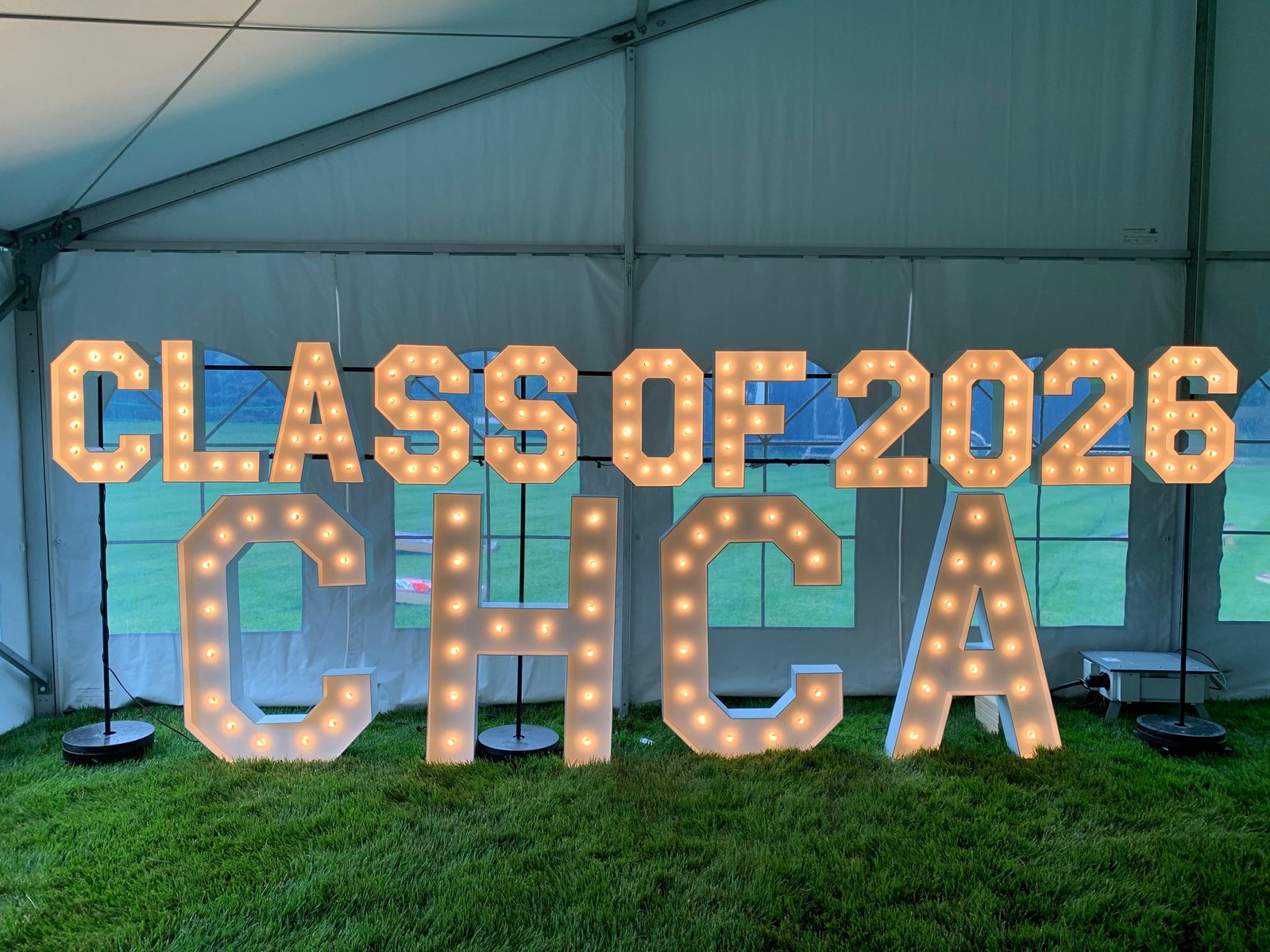 under a white outdoor event tent resting on green grass are large light up marquee letters stacked atop each other for a graduation party