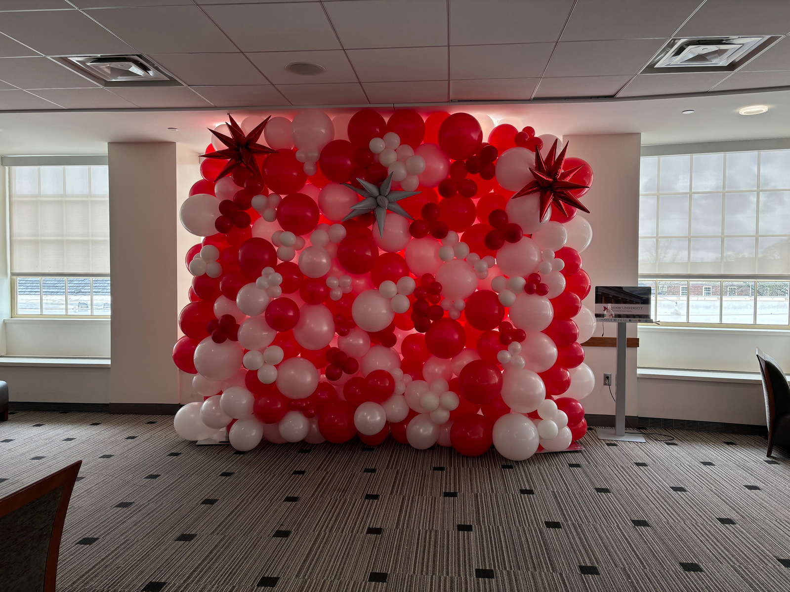 In a dim room near windows is a large balloon wall of red and white balloons with red and grey balloon spikes