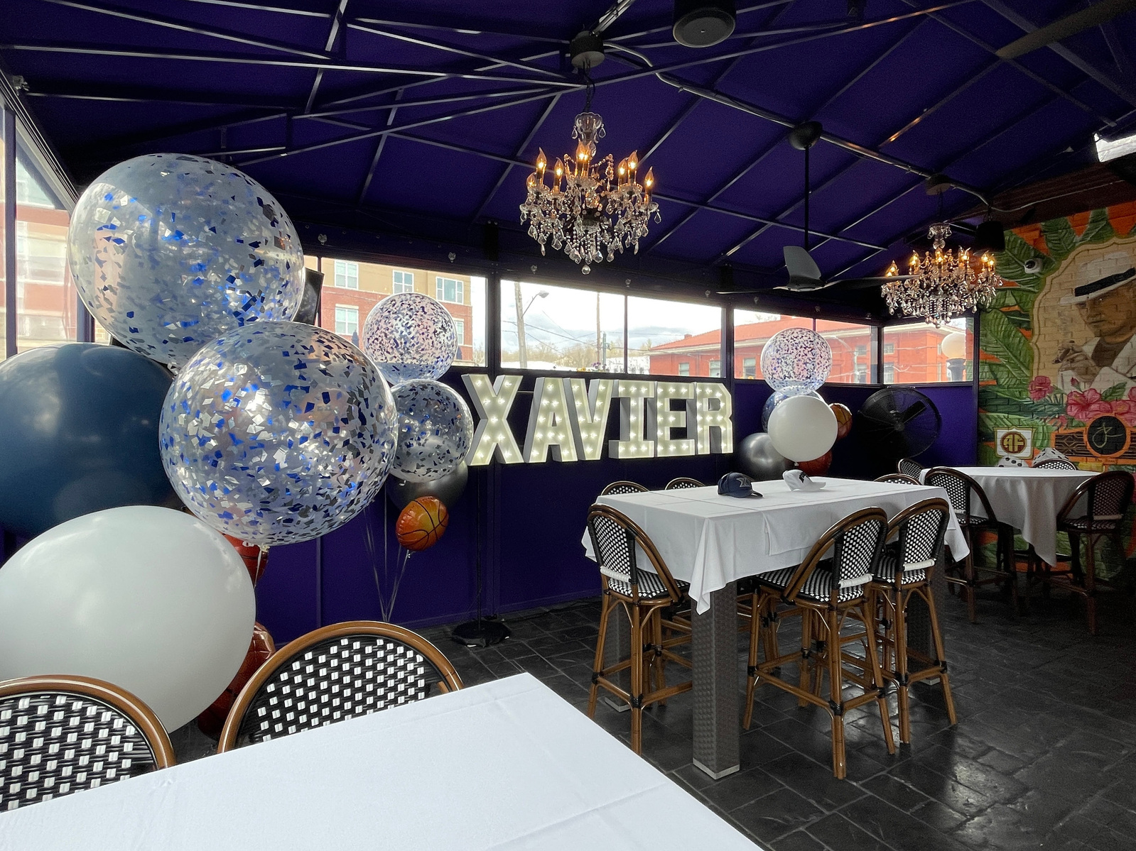 Under a purple outdoor event tent are light up marquee letters with helium balloon bouquets of blue white and clear confetti balloons