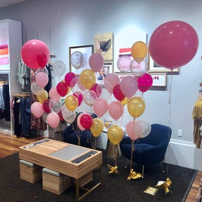 in a sitting room between a coffee table and chairs are helium balloon bouquets of pink clear and gold balloons with a large jumbo pink balloon on each side