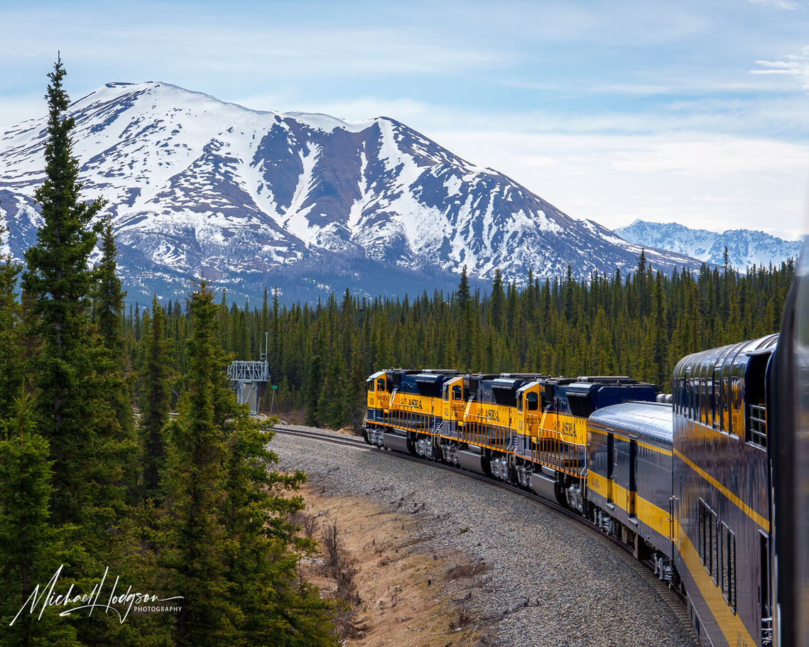Photo of an Alaska Railroad train on a curve in the track with a snow-covered mountain behind. 