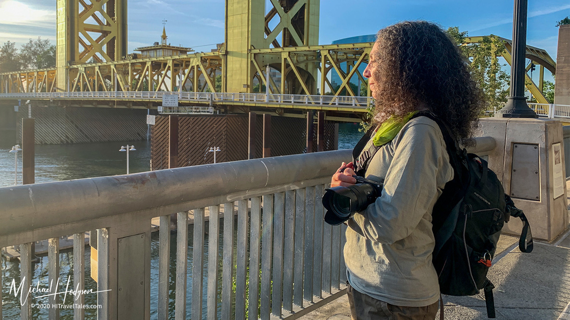 A photographer standing on the Sacramento Bridge, camera held at her side ready to take a photo without being noticed. 