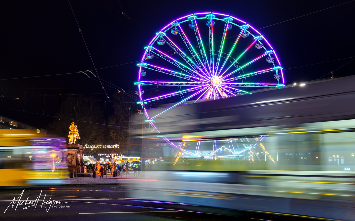 Photo of blurred street cars and trams in  Dresden as they pass in front of an illuminated Ferris wheel at night.