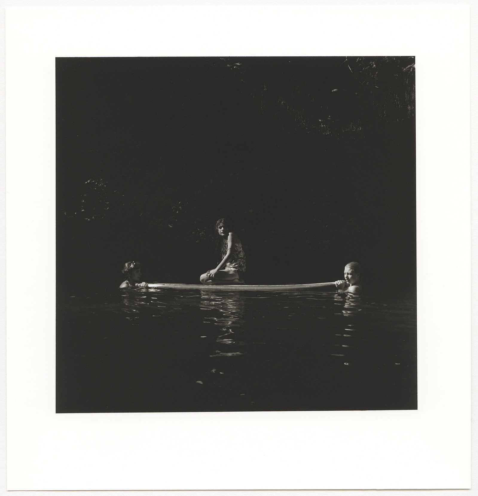 Three girls surrounded by darkness in a river, one of them is sitting on a surfboard