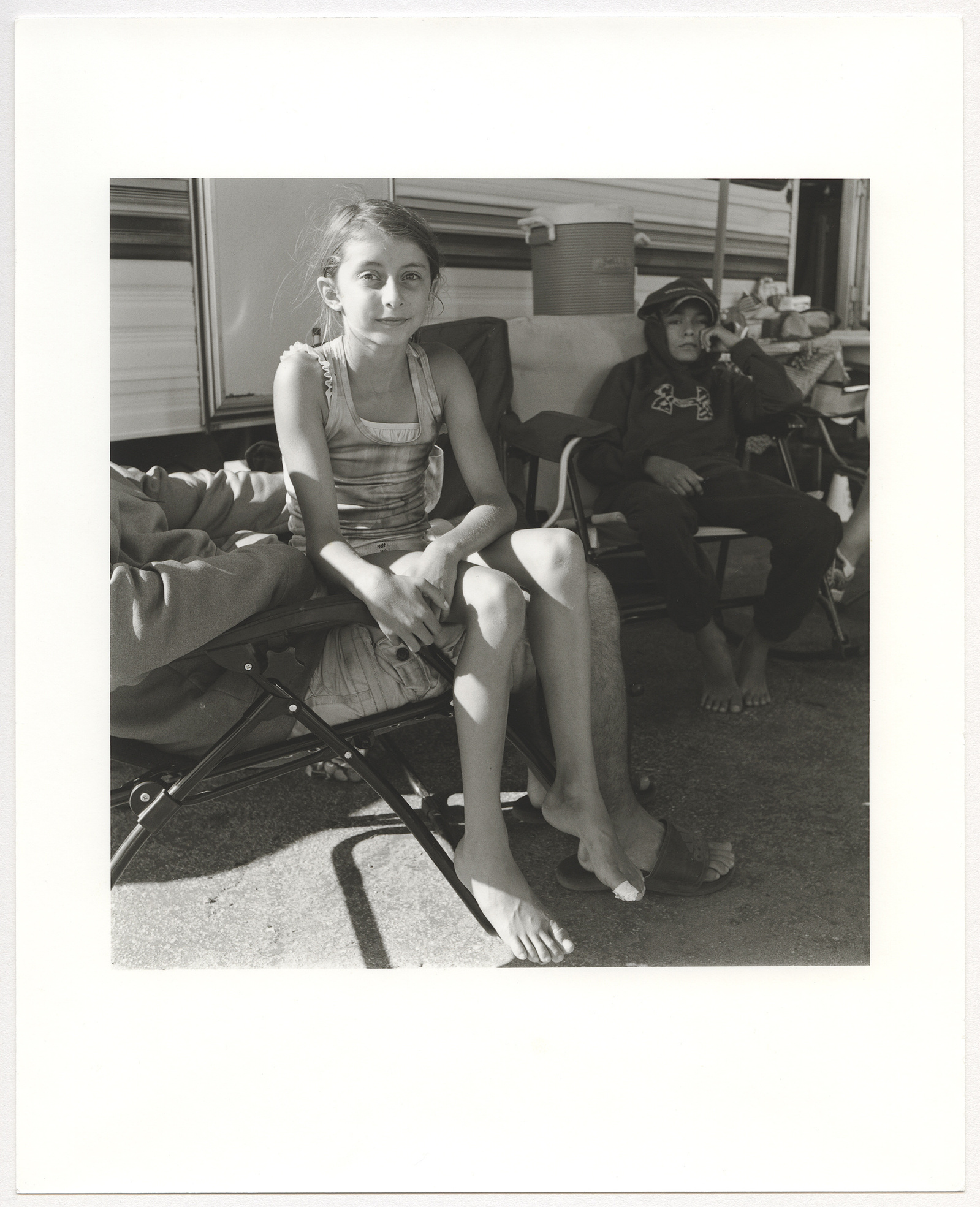 Black and white photo of a family near their RV. A girl sits on her father's lap with a bandaged toe.