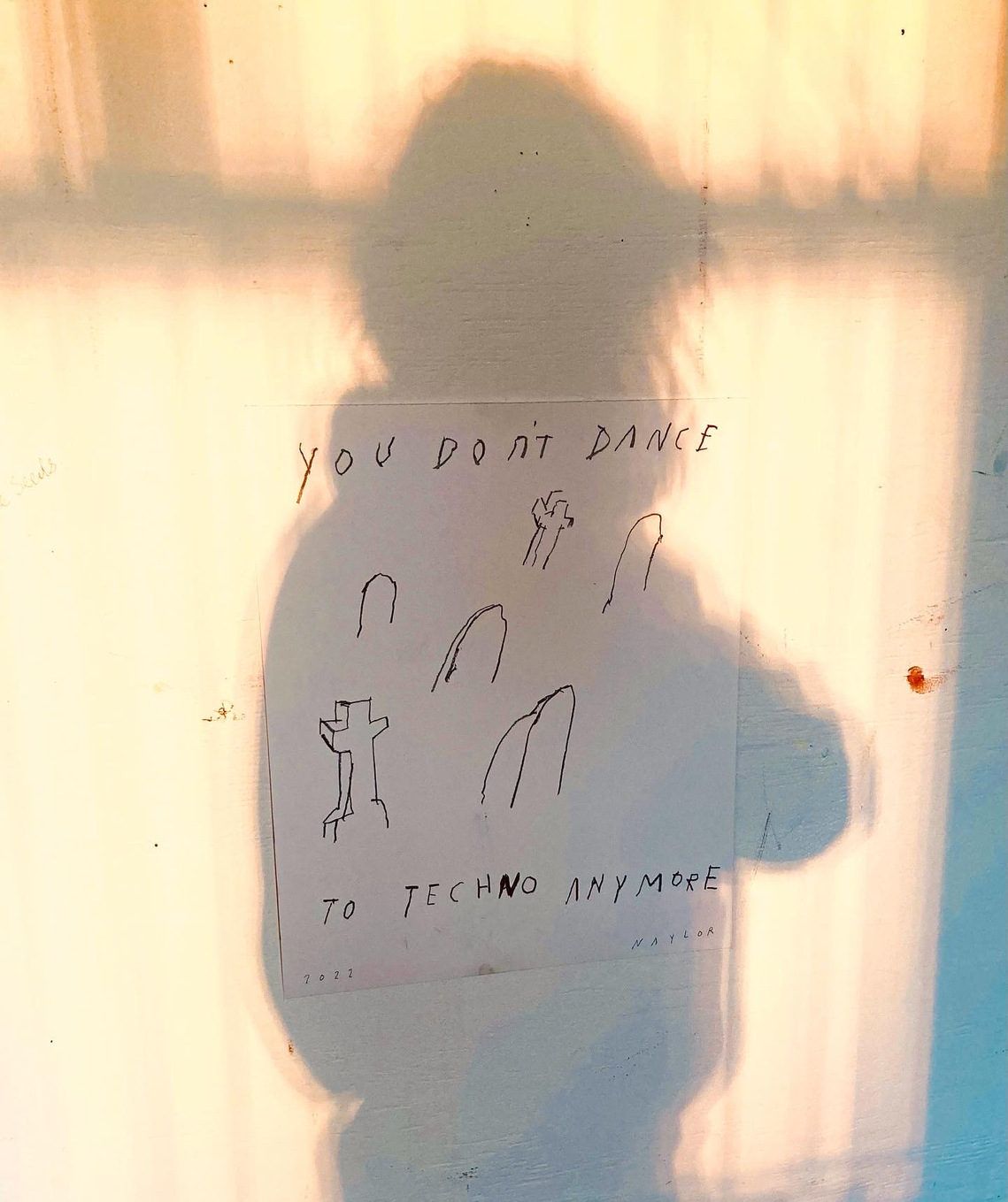 A photograph of a charcoal drawing made by the artist Mary Naylor it features graveyard with text displayed on a wall framed within a shadow of a girl. With bright light flooding in.