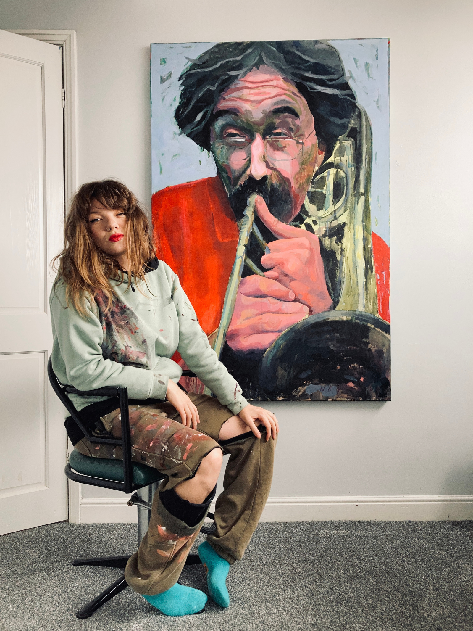Ebor Studio Artist Mary Naylor sits on a chair in front of her giant painting of the late, French, Jazz Trombonist Alain Gibert.
He wears a scorching red shirt and is set against a pale sky-blue background. She wears paint-spattered ripped clothes. 