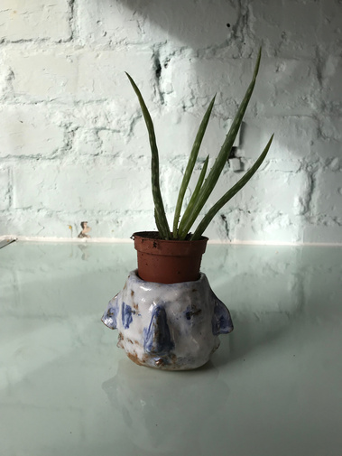 Small ceramic pot made by Mary Naylor is holding a small aloe Vera. It is blue and white and has with four connected faces around the perimeter each blending into the next. It sits on top of a glassy ice blue worktop in front of a blue painted brick wall.