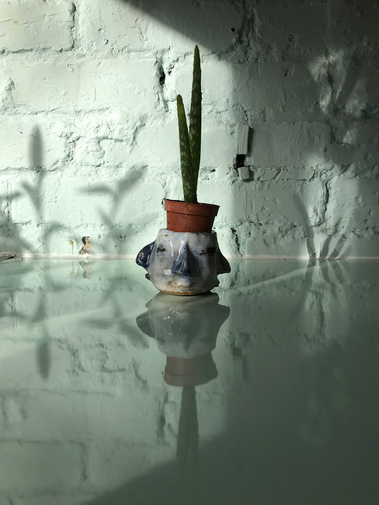 Small ceramic pot made by Mary Naylor is holding a small aloe Vera. It is blue and white and has with four connected faces around the perimeter each blending into the next. It sits on top of a glassy ice blue worktop in front of a blue painted brick wall.