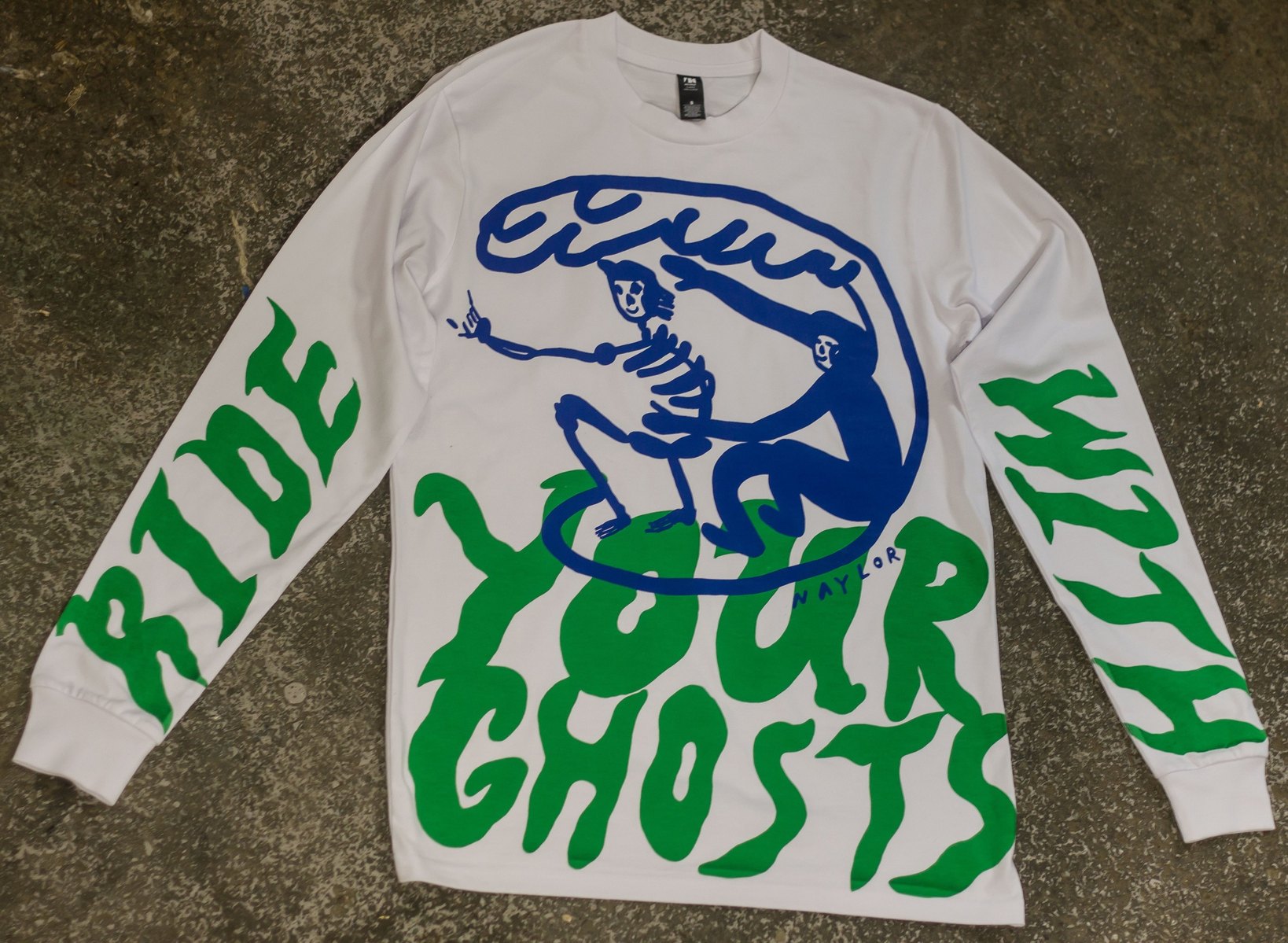 A white long-sleeve illustrated T-shirt. On the front is a blue surfer and a skeleton surfer sharing a board and a large barrel wave. There is large writing on both sleeves and the front which reads 'ride with your ghosts' This writing is green.