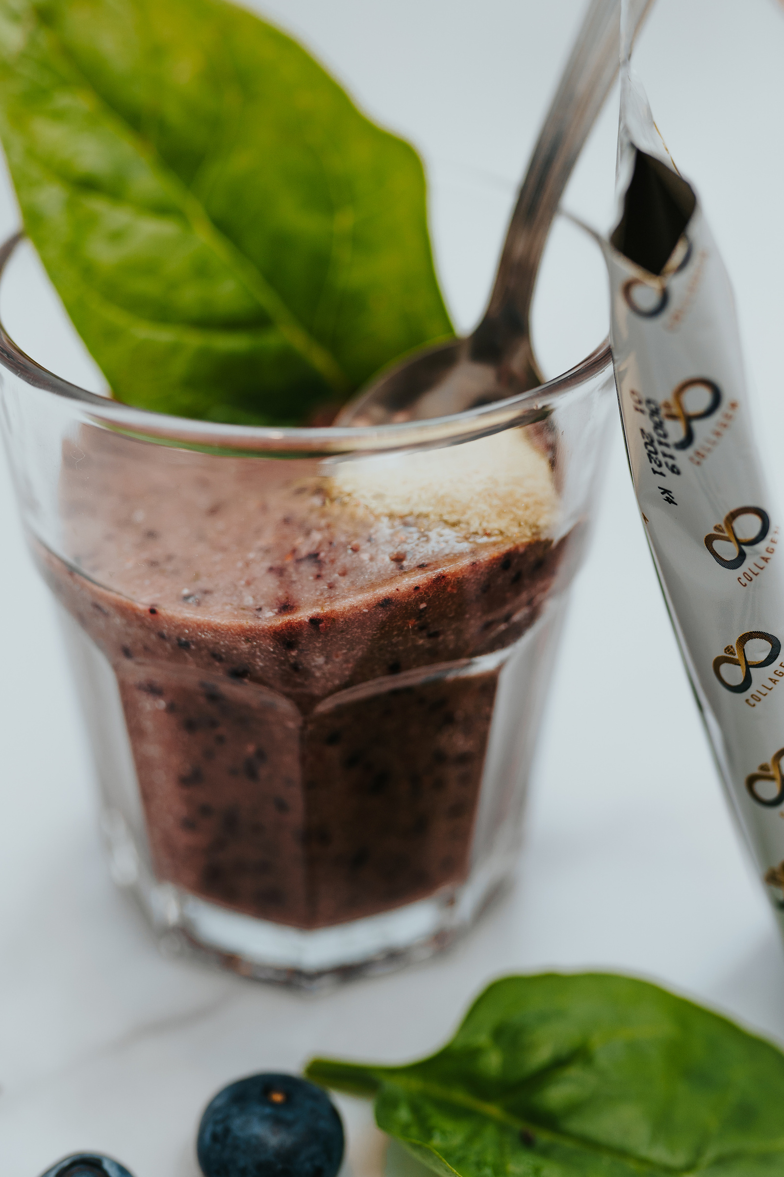 Product photography. Collagem sachet being poured into a berry smoothie and stirred with a silver spoon. Blueberries and spinach decorate the image.