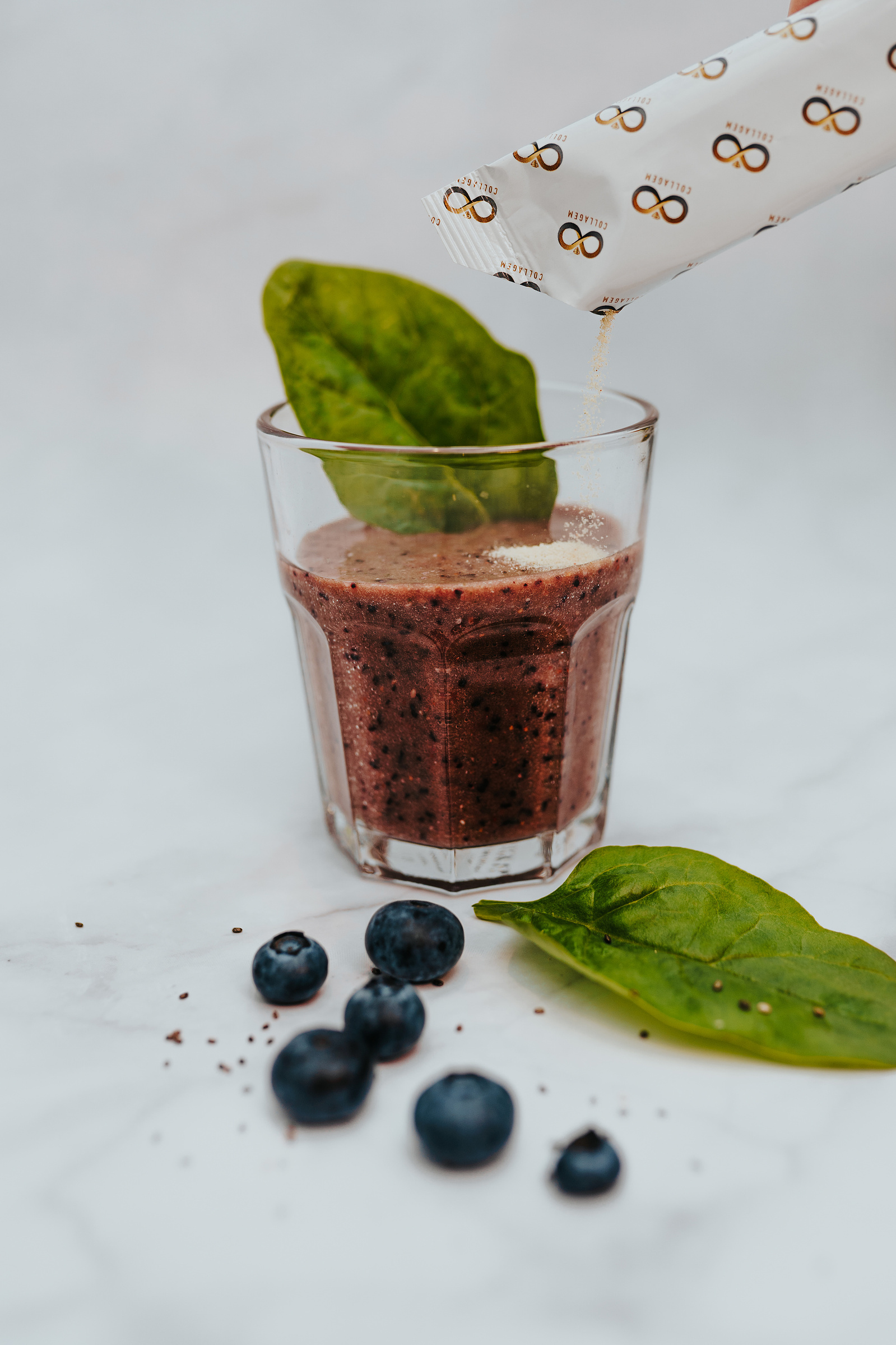 Product photography. Collagem sachet being poured into a berry smoothie. Blueberries and spinach decorate the image.