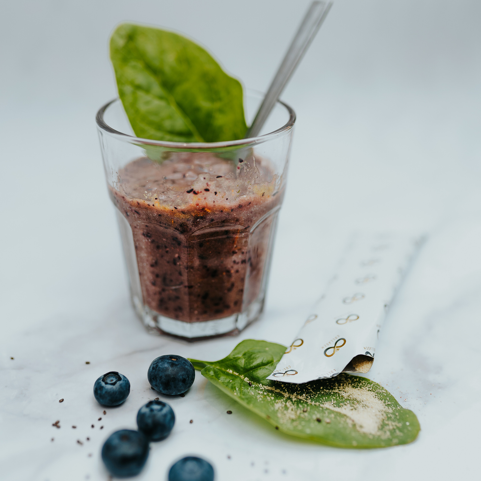 Product photography. Collagem sachet being poured into a berry smoothie. Blueberries and spinach decorate the image.