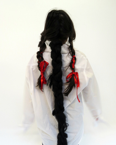 A back view of a woman in white men's shirt with black braids tied with a red ribbon, an artificial long braid attached in the centre partition of hair.