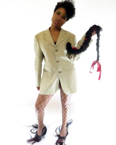 A woman from Dominic Republic in short curly hair, wearing beige men's jacket and netted stockings. Holding an artificial long black braid tied with red ribbon. Shoes have fake hair all over.