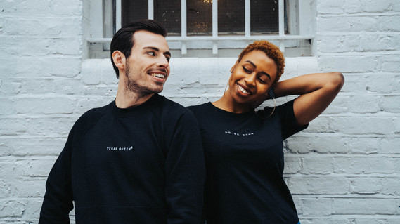 Two models smiling at each other in front of a white wall at a fashion photoshoot.