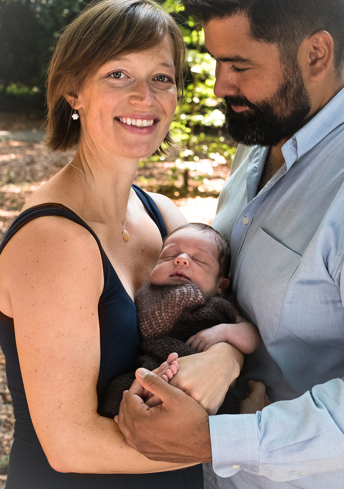 Face to face, dad gazes at mum who smiles to camera, they are both holding their newborn son in their arms. Family and newborn photographer, Chene-Bougeries.