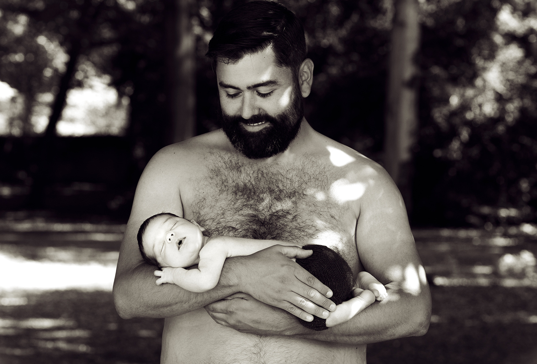 Black and white portrait of a bare-chested young loving father, holding his first baby in his arms. Family and newborn photographer, Helen Putsman, works from Chene-Bougeries, Geneva.