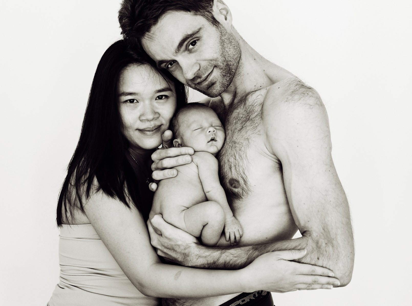 Loving parents pose for a family portrait, the father holds his newborn son close to his bare chest for their first professional portrait as a family. Photographed by Geneva's leading portrait photographer, Helen Putsman, based in Chêne-Bougeries.