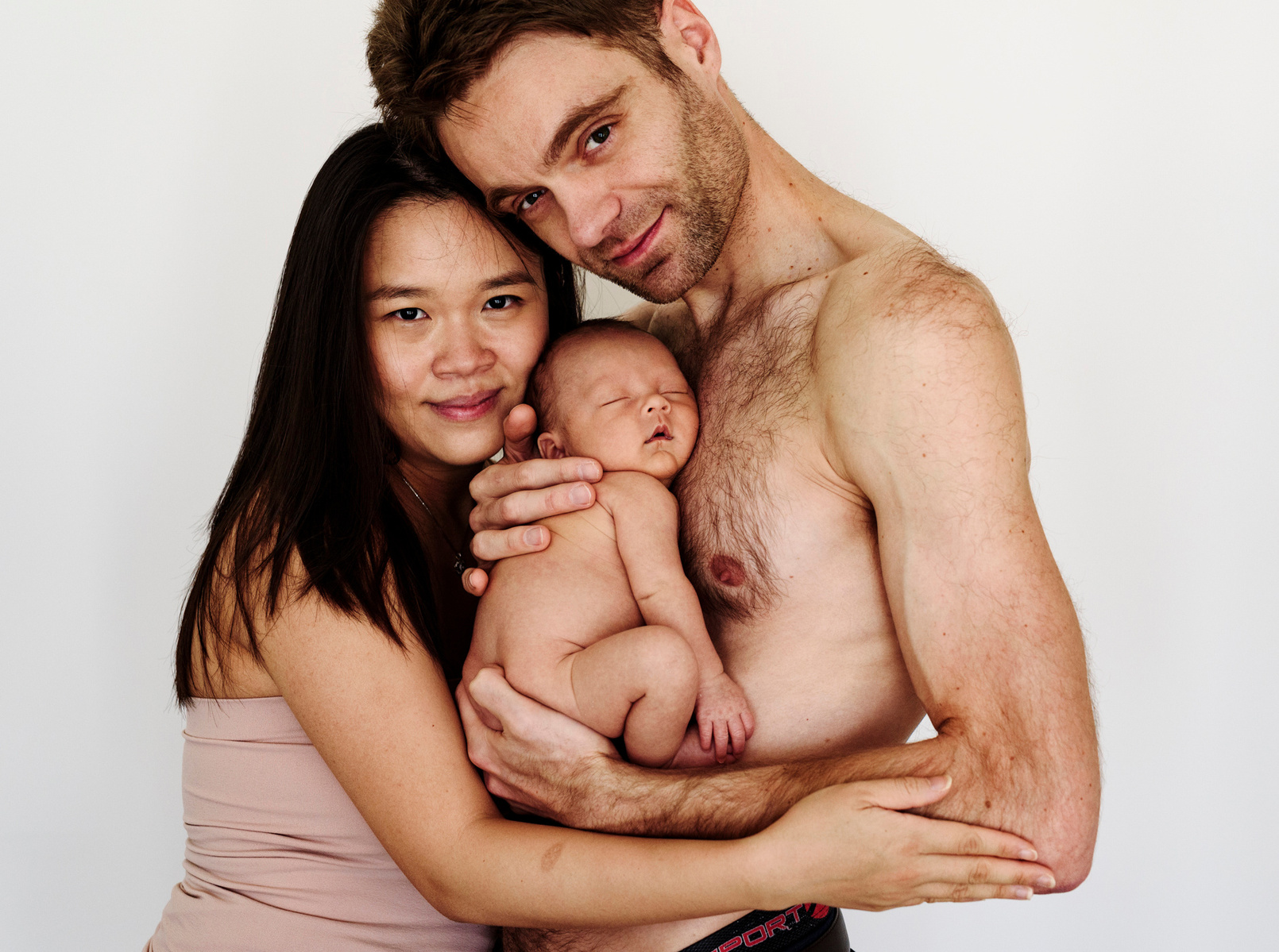 Loving parents pose for a family portrait, the father holds his newborn son close to his bare chest for their first professional portrait as a family. Photographed by Geneva's leading portrait photographer, Helen Putsman, based in Chêne-Bougeries.
