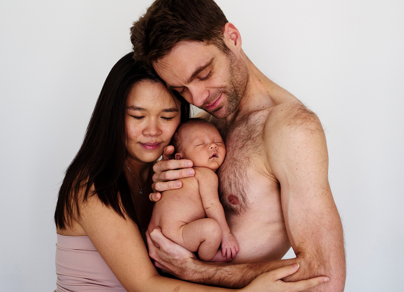 Loving parents pose for a family portrait, the father holds his newborn son close to his bare chest for their first professional portrait as a family. Photographed by Geneva's leading portrait photographer, Helen Putsman, based in Chêne-Bougeries.