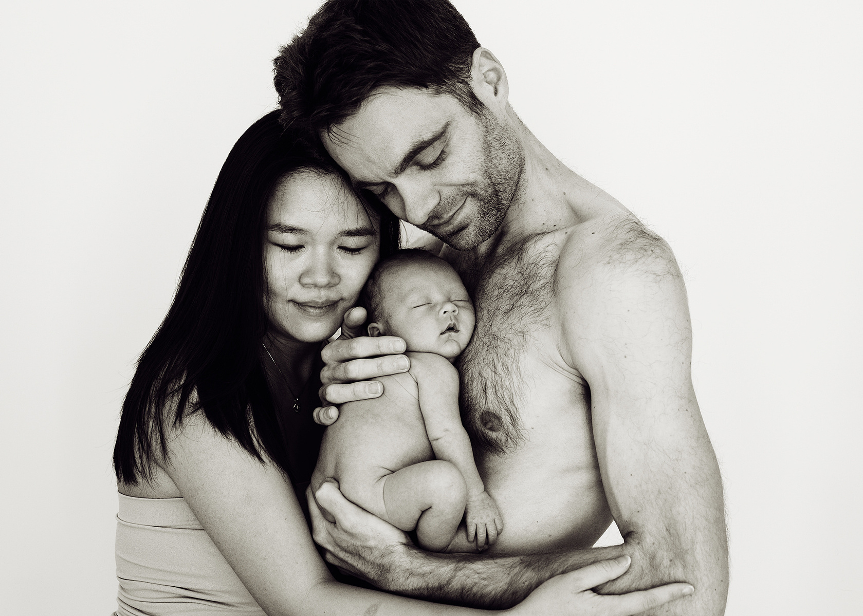 Loving parents pose for a family portrait, the father holds his newborn son close to his bare chest for their first professional portrait as a family. Photographed by Geneva's leading portrait photographer, Helen Putsman, based in Chêne-Bougeries.