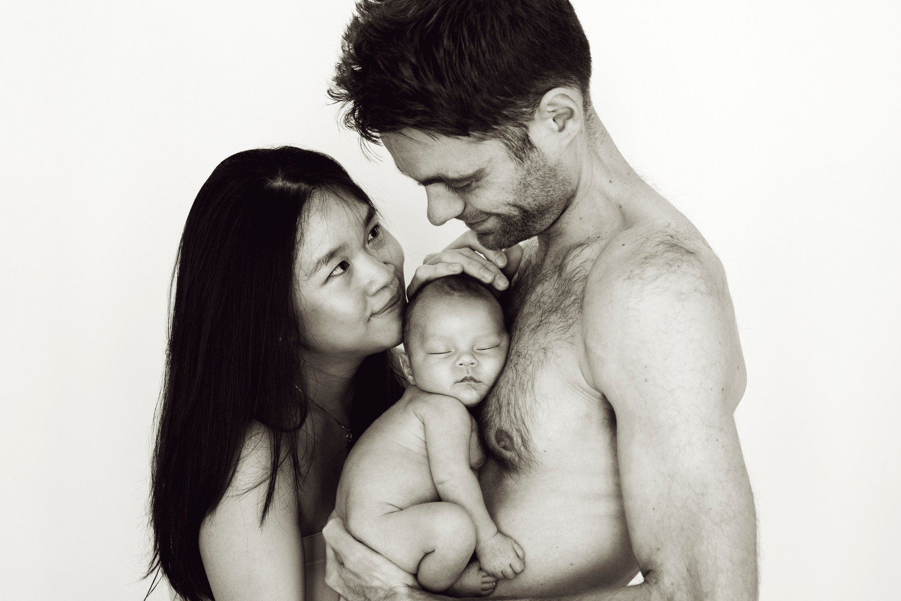 Loving parents look into each other's eyes whilst the father holds his newborn son close to his bare chest for their first professional portrait as a family. Photographed by Geneva's leading portrait photographer, Helen Putsman, based in Chêne-Bougeries.