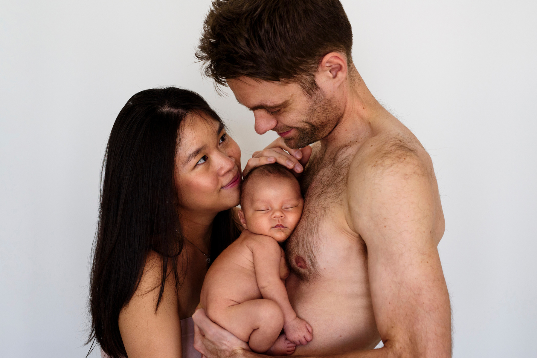 Loving parents look into each other's eyes whilst the father holds his newborn son close to his bare chest for their first professional portrait as a family. Photographed by Geneva's leading portrait photographer, Helen Putsman, based in Chêne-Bougeries.