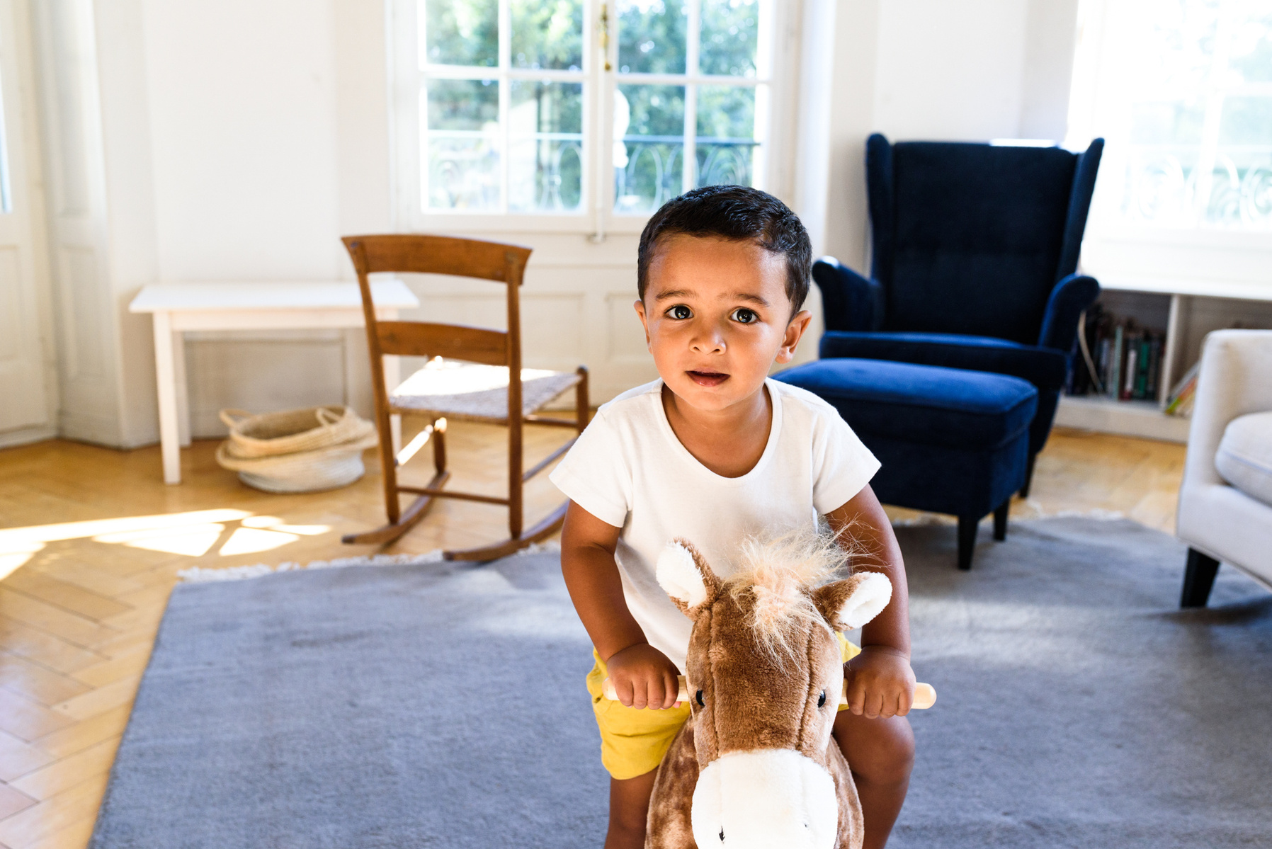 Lifestyle photography of a little boy playing on his rocking horse in his family's lounge with the sun streaming in. Shot in Geneva, Switzerland.
