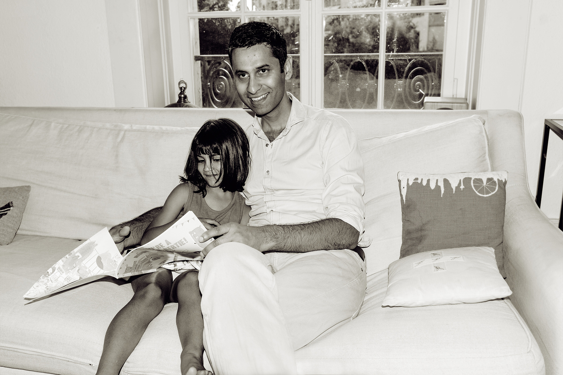 Father looks at the camera for a portrait with his daughter cuddled up closely to him whilst he reads her a story. Shot in Geneva, Switzerland.