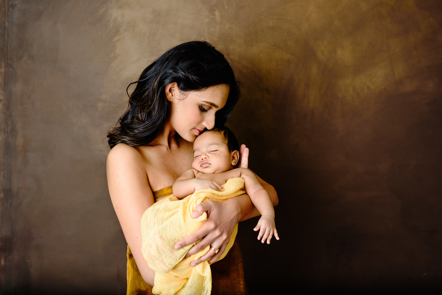 Photograph of a young mother holding her wrapped newborn son in her arms, set on a brown hand-painted canvas. This portrait was shot during a newborn photoshoot, available here: https://www.helenputsman.com/