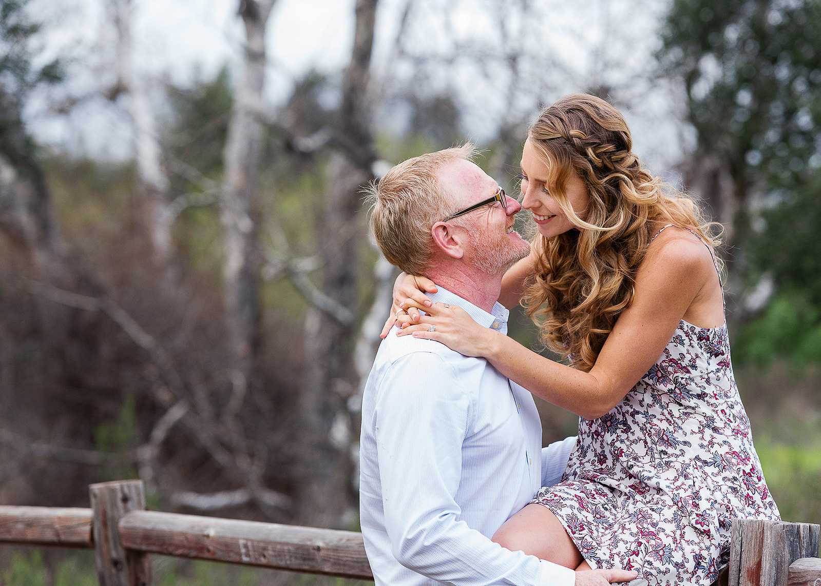 Couple poses for engagement session on fence with trees in the background.