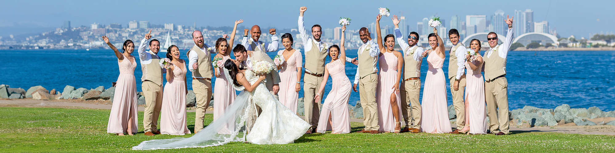 Bride and groom kissing with wedding party celebrating taken from Shelter Island with the San Diego skyline in the background.