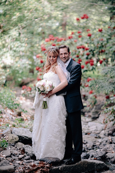 Bride and groom standing on rocks in creek at Arnold Arboretum, surrounded by trees with red flowers.