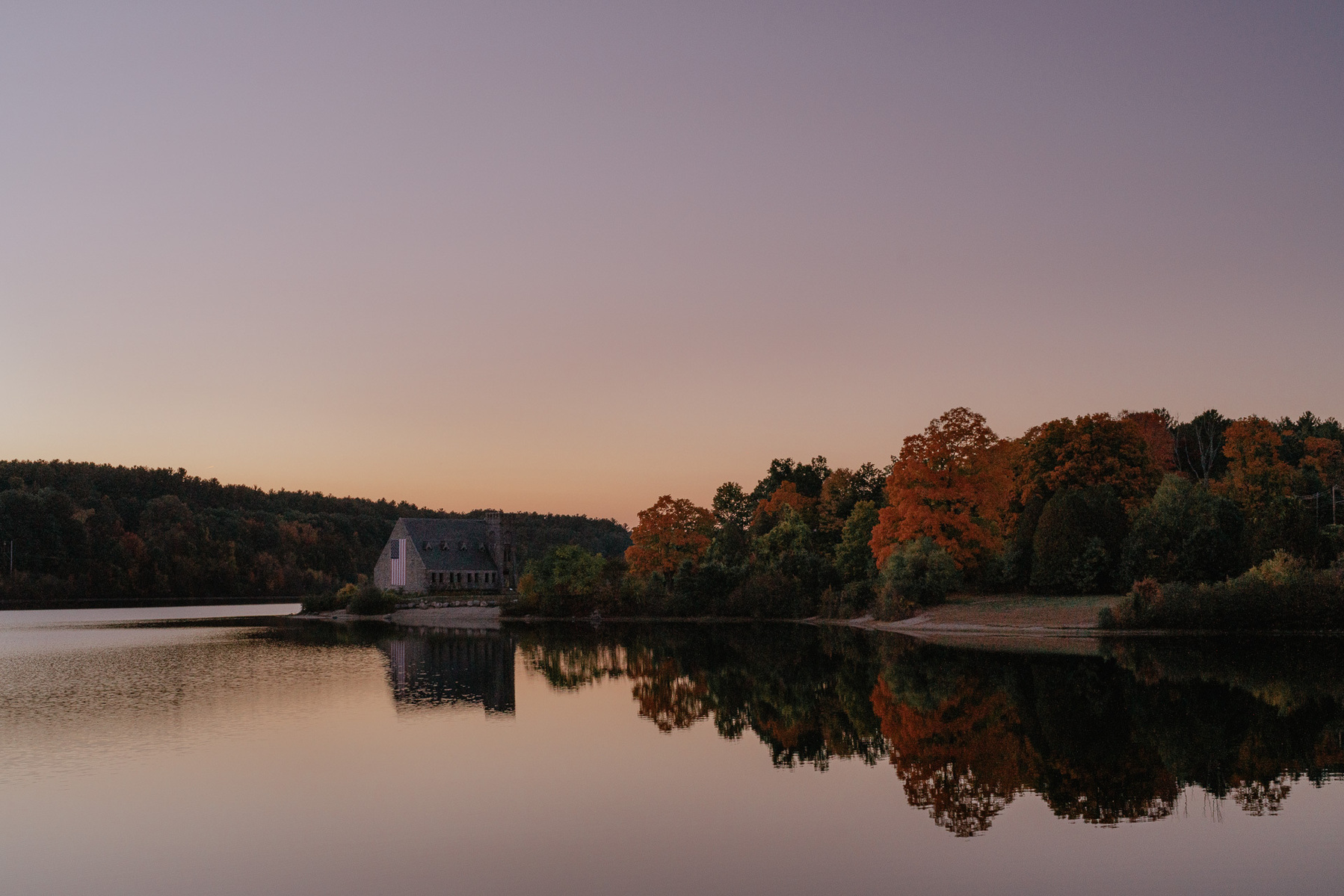 Old Stone Church in West Boylston, MA, reflecting in lake during sunset with fall foliage.