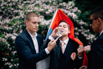 Groomsman making groom drink in front of Serbian flag.