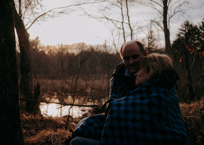 Older couple cuddling under blanket by river at sunset.