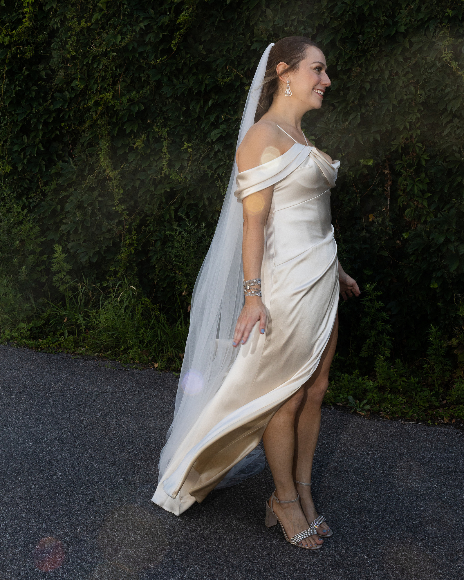 Bride in wedding dress and veil stands in front of greenery looking right. Wedding photojournalism at Senate Garage in Kingston, NY.