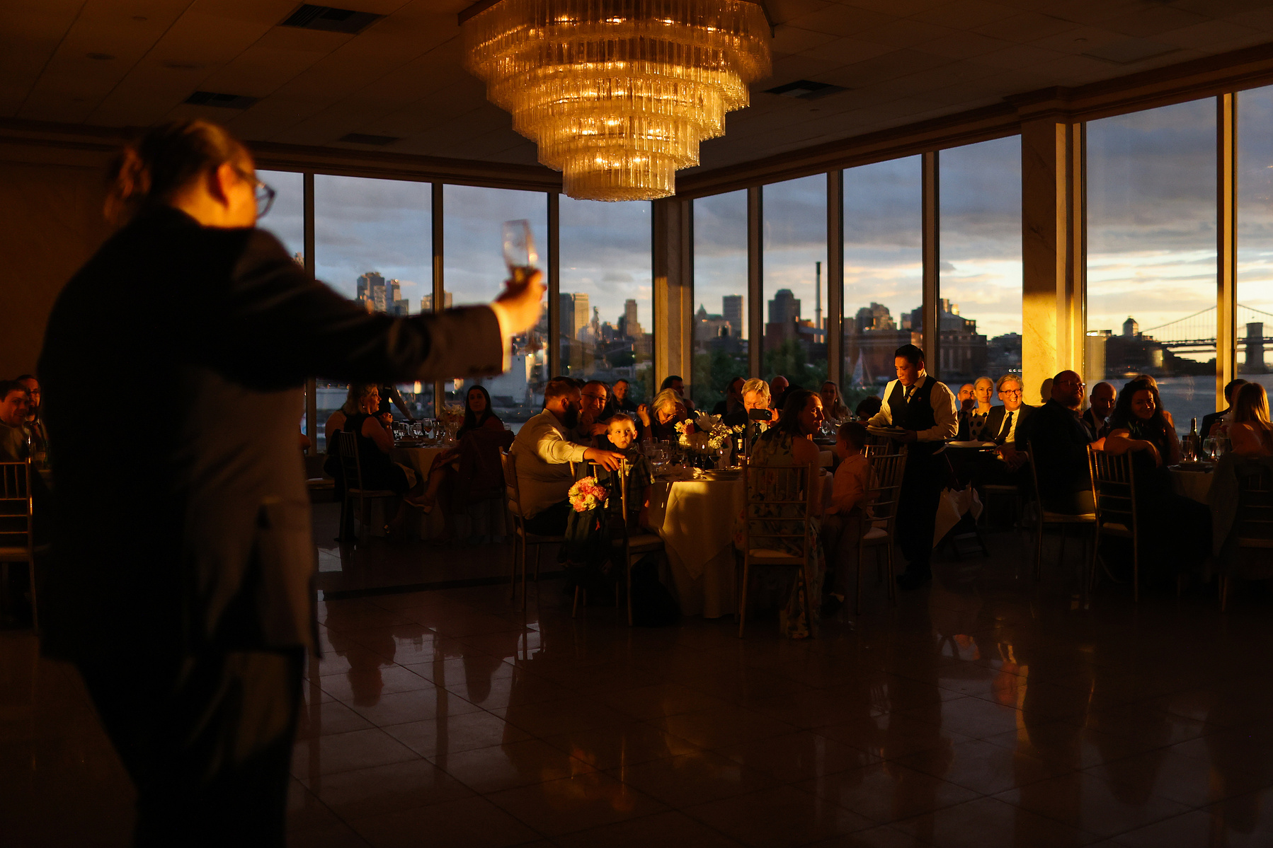 Groom gives toast to room bathed in orange light from the setting sun. Wedding photojournalism at Giando on the Water in Brooklyn, NY. 