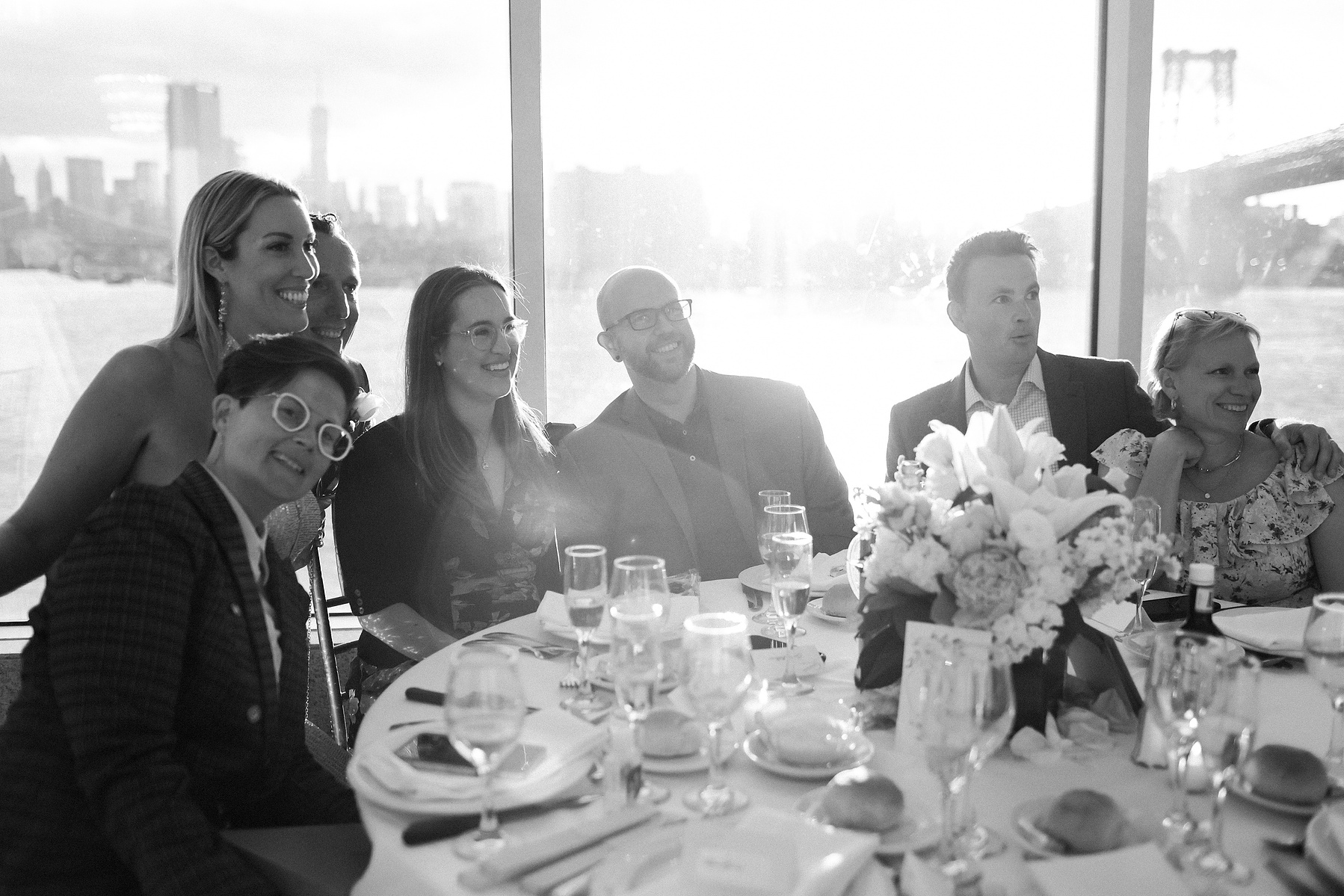Wedding guests at reception table as strong directional light from the setting sun streams in. Wedding photojournalism at Giando on the Water in Brooklyn, NY. 