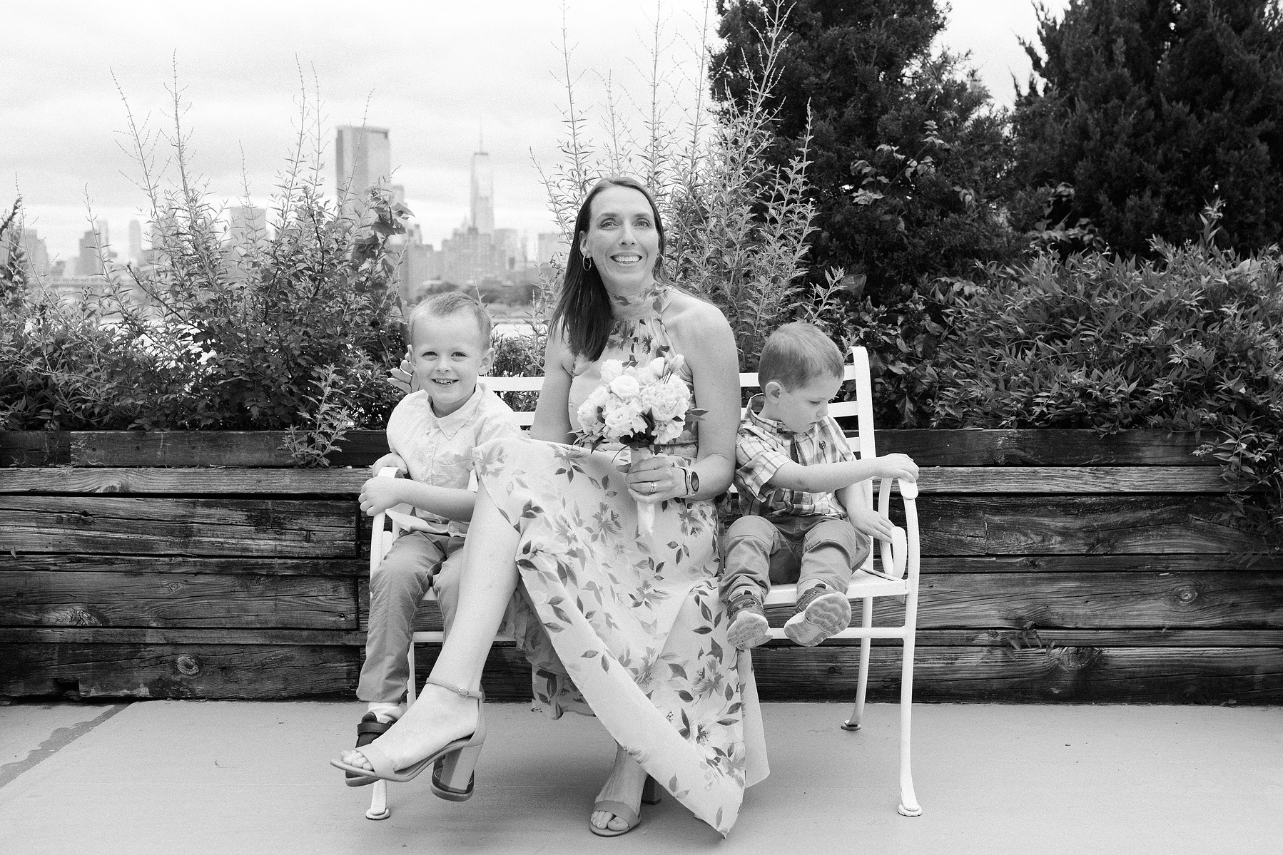 Woman sitting on bench outside by the water with her two children. Wedding photojournalism at Giando on the Water in Brooklyn, NY. 