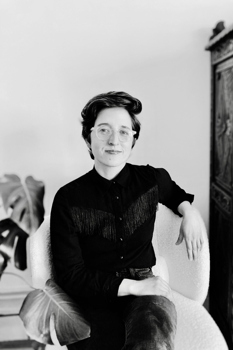 k stark sits in a white armchair in front of a white wall, their left arm draped over the armrest and their right resting on their lap. they are wearing a black fringe cowboy shirt. to the left of them is a monstera and to the right is a wood armoire. 