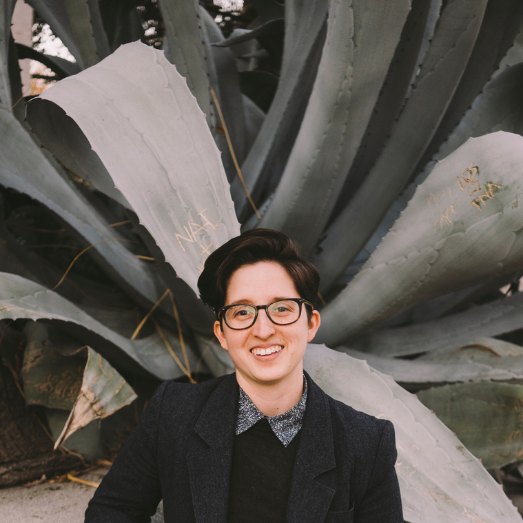 karly stands in front of a large green succulent plant, which towers over their head. they smile and look at the camera, wearing glasses, a blue button up shirt and grey blazer. 