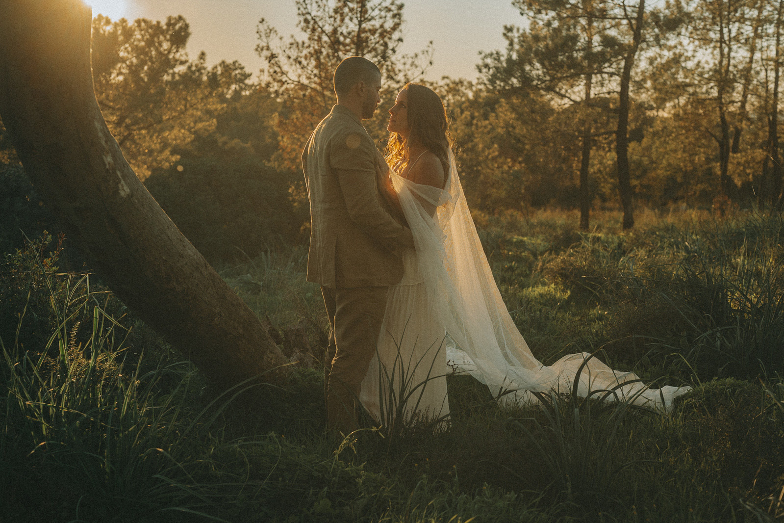 A just married couple is standing in a forest,  looking at each other in their in a white wedding dress and a casual linen suit at the Algarve in Portugal.